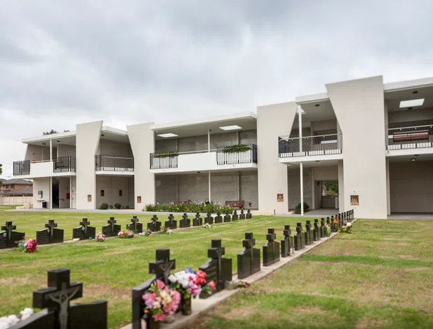 Exterior view of a contemporary multi-storey building with pale rendered walls, projecting balconies, open circulation areas, and a large covered ground-floor opening. The building overlooks a landscaped lawn with rows of small dark memorial crosses and flower arrangements. Overcast sky and restrained geometric architecture create a solemn institutional setting.