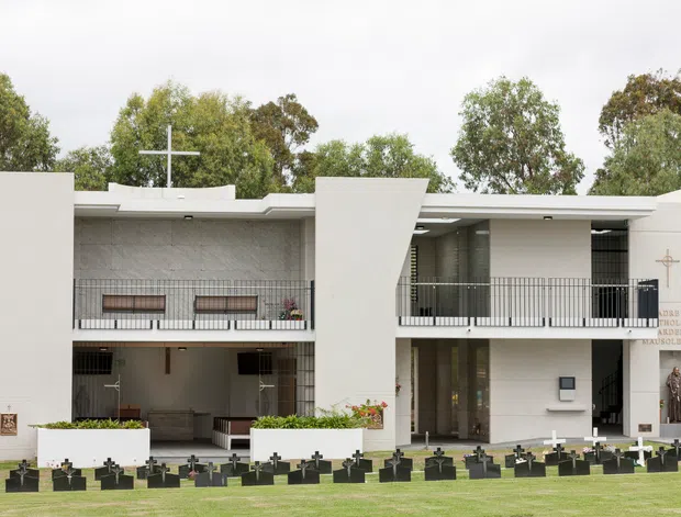 Exterior view of a modern two-storey memorial or religious building with white rendered walls, projecting geometric facade elements, open upper-level balconies, dark metal railings, and extensive glazing. Crosses and memorial plaques indicate a church, chapel, or mausoleum setting, with landscaped grounds and rows of small dark memorial markers in front.
