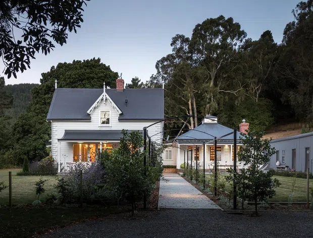 Exterior view of a white weatherboard country residence with a steep gabled roof, dormer window, verandah, and warm-lit windows. A gravel path leads through a landscaped garden to the main house, with a connected lower wing or outbuilding to the right. Dense mature trees and a wooded hillside form the background, creating a rural, heritage-inspired setting.