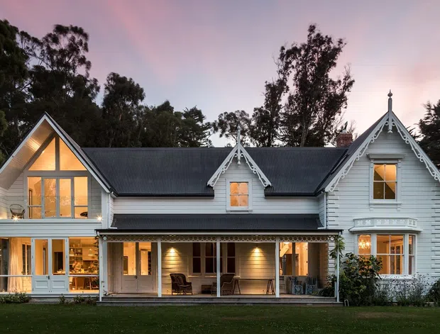 Exterior view of a substantial white weatherboard house at dusk, featuring multiple steep gabled roof sections, ornate timber gable trim, dormer windows, large glazed openings, and a covered verandah running across the ground floor. The home is set within a mature wooded landscape and has warm interior lighting visible through the windows.