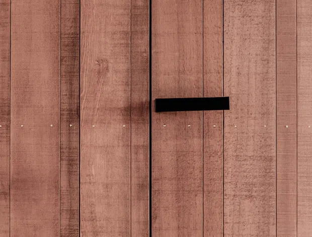 Close-up of a vertically boarded timber-clad surface in a warm reddish-brown tone, with visible wood grain, narrow dark joints, regularly spaced fasteners, and a horizontal black rectangular handle or hardware element. The image is a detail study of a flat architectural surface with no wider room context visible.