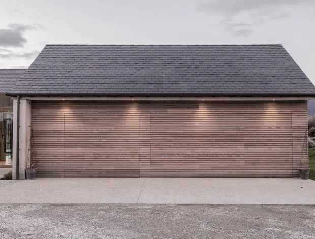 Front exterior view of a low-profile residential garage or carport structure with a broad timber-clad garage door, dark shingle-like pitched roof, and simple cylindrical support columns. The building sits on a gravel and concrete driveway in a landscaped setting, with subdued overcast lighting and distant hills visible at the edges.