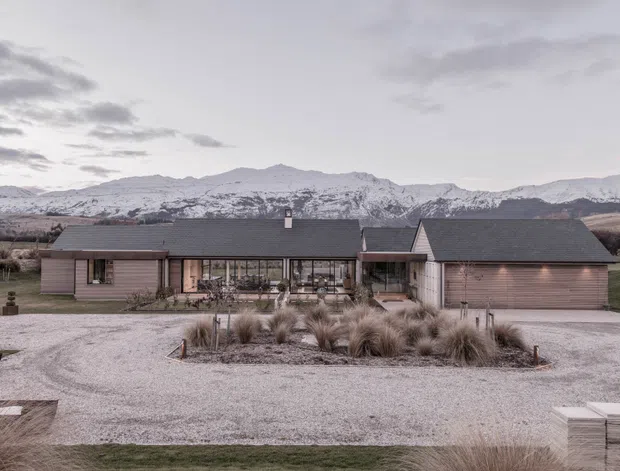 Wide exterior view of a low, sprawling residential home set against snow-covered mountains. The house has a dark pitched roof, muted timber-clad walls, extensive glazing across the central living area, and an attached garage. A pale gravel driveway and minimalist landscaped planting sit in the foreground.