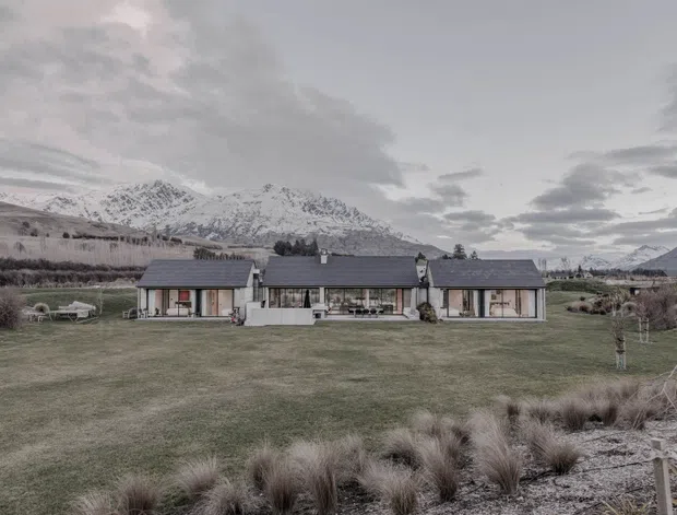 A low, single-storey modern residence arranged as three connected pitched-roof volumes, set in an expansive landscaped lawn with dramatic snow-capped mountains behind it. The dark grey roof planes, restrained neutral exterior finishes, large glazed openings, and central covered outdoor area create a contemporary alpine composition. The image is presented in a muted, overcast palette.