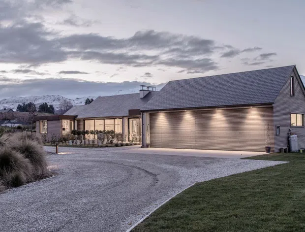 Twilight exterior view of a low, single-storey residential home with a broad gabled roof, attached double garage, muted grey horizontal cladding, and warm exterior lighting. A gravel driveway and landscaped grasses lead toward the house, with snow-dusted mountains visible in the background.
