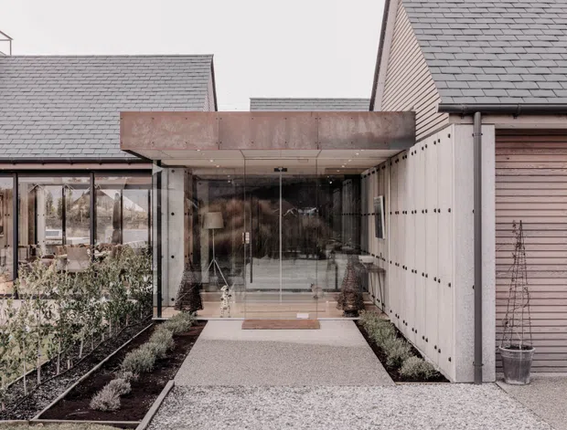 Exterior view of a contemporary residential entrance linking two pitched-roof volumes with a glazed passage. The composition features a projecting weathered metal canopy, extensive floor-to-ceiling glazing, pale vertical cladding, timber-toned horizontal siding, and a gravel forecourt with structured planting beds.