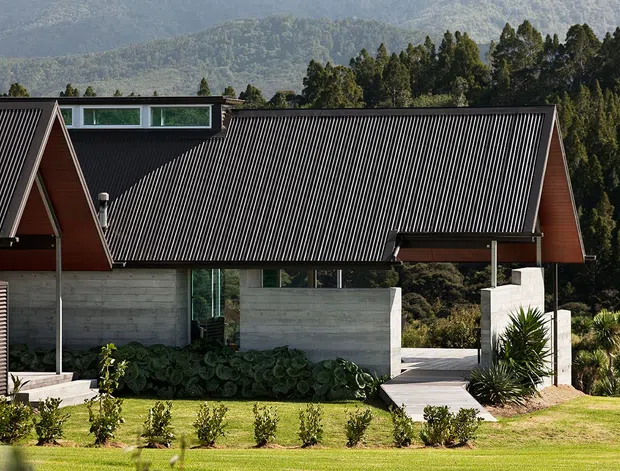 Exterior view of a contemporary single-storey house set against a wooded mountain backdrop. The building has steep gabled forms, dark ribbed metal roofing, reddish timber-clad gable walls, exposed concrete walls, clerestory glazing, and a landscaped lawn with a paved walkway.