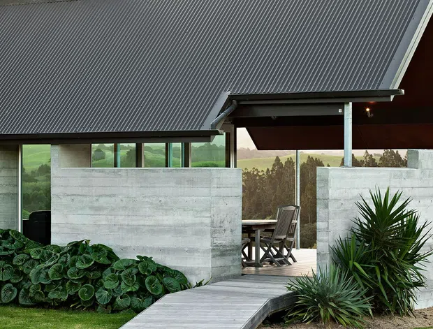 Exterior architectural view of a contemporary single-storey home with a steeply pitched corrugated metal roof, exposed dark timber soffits, concrete block walls, extensive horizontal glazing, and a covered outdoor dining terrace. A timber walkway leads through landscaped planting toward the sheltered patio.