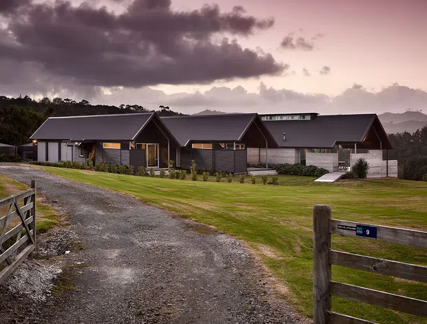Exterior view of a contemporary single-storey rural residence composed of several connected dark-clad gabled wings. The home sits beyond a gravel driveway and timber farm-style gate, with open lawns, landscaped planting, surrounding trees, and distant hills beneath a dramatic cloudy sky.