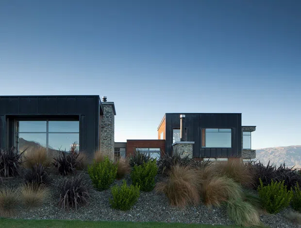 A low-profile contemporary residence composed of dark rectangular volumes with large recessed glazing, pale stone chimney or wall elements, and warm timber detailing. The house is set into a landscaped, dry-climate garden with ornamental grasses and shrubs, overlooking a broad mountainous landscape under a clear blue sky.