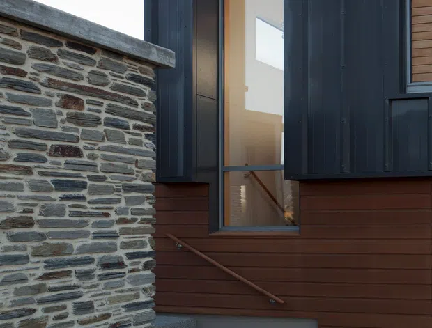 Exterior architectural view of a contemporary multi-level building, focused on a stairway rising beside a low rough-stone masonry wall. The facade combines dark vertically ribbed metal cladding, warm horizontal timber boards, and large glazed openings revealing an interior stair. The composition has a restrained, material-focused palette and strong geometric lines.