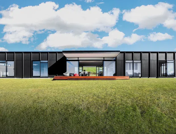 A low, elongated contemporary home set in an open grassy landscape. The facade is composed of dark vertical cladding panels and extensive floor-to-ceiling glazing, with a recessed central outdoor living area and a broad projecting roof canopy. The image is an exterior architectural view under a bright blue sky.