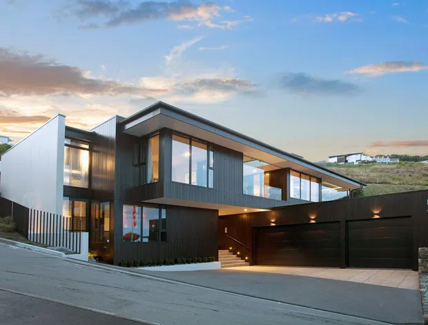A contemporary multi-level residence on a sloping suburban site, viewed from the street at dusk. The home features dark vertical cladding, expansive glazing, crisp cantilevered roof planes, an integrated double garage, and a landscaped entry stair. A lighter rendered wall and neighbouring homes are visible in the background.