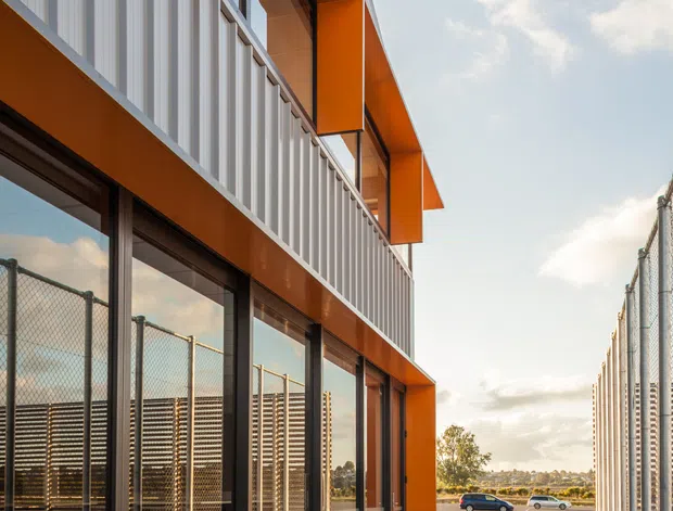 Exterior view of a contemporary multi-storey commercial or institutional building with bold orange projecting frames, dark metal-framed floor-to-ceiling glazing, and vertically ribbed light-grey facade panels. A wide concrete walkway runs alongside chain-link fencing, with an open paved area and distant landscape beyond.