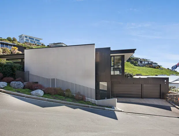 Street-level exterior view of a contemporary multi-level residence on a sloping suburban site. The house features a flat roof, crisp rendered walls, dark vertical cladding, large windows, an integrated double garage, and minimalist landscaped planting with boulders and grasses. Adjacent hillside homes and an Australian flag are visible in the background.