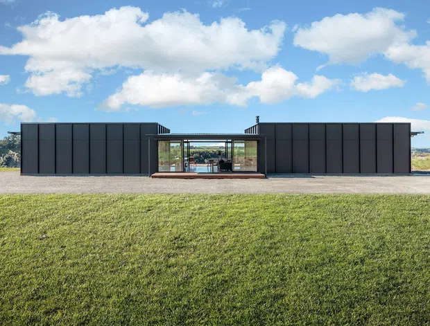 A low, single-storey modern building viewed straight on across a broad lawn. Two long, dark charcoal-clad wings are connected by a recessed glazed central entrance or courtyard, creating a strongly symmetrical horizontal composition. The flat-roofed structure has large expanses of dark vertical-panel cladding and extensive central glazing, set in an open rural or semi-rural landscape beneath a bright blue sky.