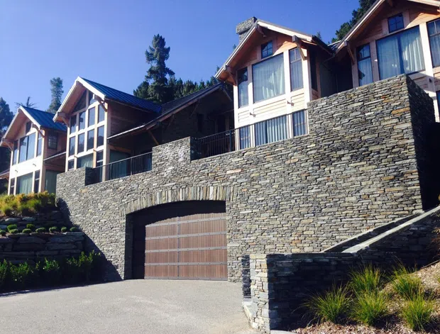 Exterior view of a multi-level mountain-style residence built into a sloping site. The home features extensive stacked-stone retaining walls and facade elements, timber-framed gabled upper volumes, dark metal pitched roofs, large glazed windows, balconies, and an integrated double garage.