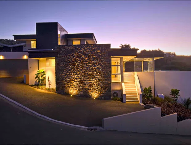 Twilight exterior view of a contemporary multi-level residence on a sloping site. The home features a flat-roofed composition, dark rendered and stone-clad volumes, warm accent lighting, a garage, glazed entry stair, and landscaped retaining walls.