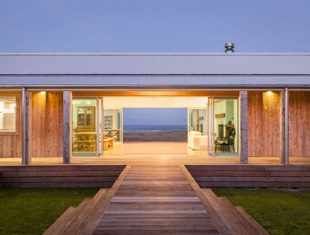 Single-storey coastal house viewed from the landscaped approach, with a broad corrugated metal roof, timber-clad facade, extensive sliding glass doors, and a central timber walkway leading into a warmly lit open-plan interior. The ocean is visible through the central opening, while built-in outdoor timber seating frames the entrance.