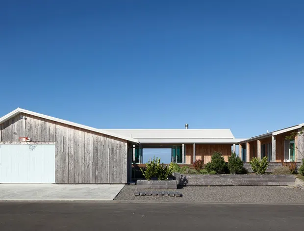Single-storey residential home viewed from the street, composed of weathered timber-clad gabled wings connected by a lower central glazed living area. The facade includes a large white garage door, timber-framed windows, a low-pitched metal roof, and a simple gravel and paved forecourt with sparse landscaping. Clear blue sky and distant landscape are visible beyond the property.