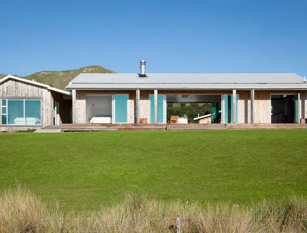 Single-storey contemporary residential building set in a broad grassy landscape with low hills behind it. The house has a long, low horizontal form, a simple shallow pitched metal roof, extensive glazed openings, timber-clad walls, and a covered outdoor veranda or deck. A smaller projecting wing sits to the left, and a chimney is visible near the roof centre.