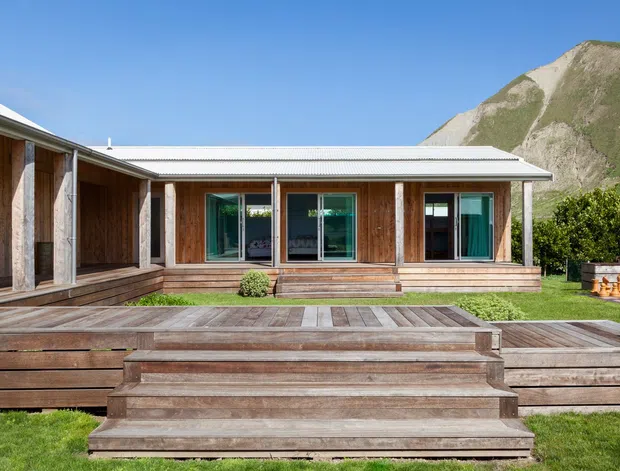 Single-storey timber-clad house viewed from a broad timber deck and stepped platform. The building has a low-pitched corrugated metal roof, large glazed openings with teal-toned frames, and a courtyard-like outdoor area. A steep grassy and rocky mountain slope rises directly behind the home.