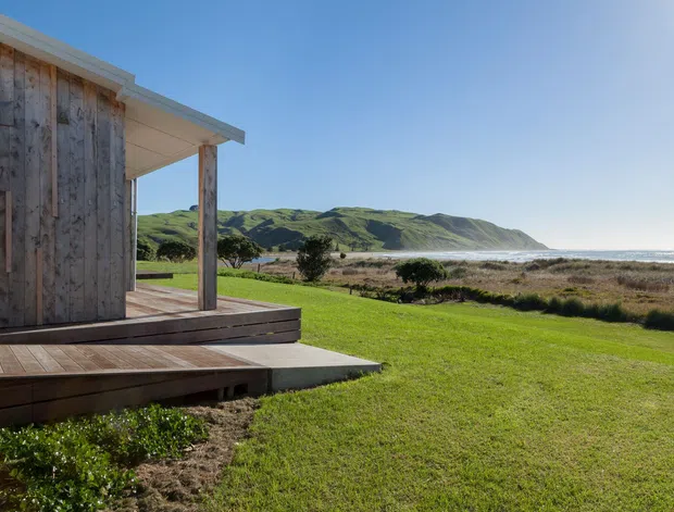 Exterior view of a compact timber-clad pavilion or cabin with a raised timber deck, set in a grassy coastal landscape overlooking dunes, surf, and rolling hills. The image highlights the building's weathered vertical timber walls, simple roof overhang, outdoor deck, and strong connection to the surrounding coastline.
