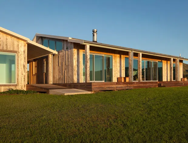 Single-storey contemporary residential building with a long, low-profile form, extensive floor-to-ceiling glazing, weathered timber cladding, and a broad timber deck. The house sits in an open grassy setting beneath a clear sky, with a visible metal flue and simple projecting rooflines.