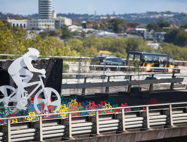 Urban exterior view of a bridge or elevated transport structure featuring a large white cyclist sculpture in the foreground, colourful bicycle-themed graphics along the barrier, metal guardrails, and construction or transit infrastructure. A cityscape is visible in the background.