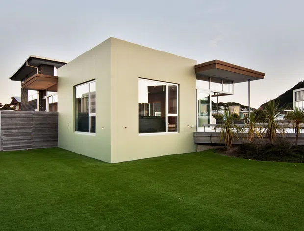 Exterior view of a modern multi-level residential property with a pale rendered central volume, extensive glazing, projecting flat roof canopies, dark timber balcony and privacy fencing, and a landscaped lawn with architectural planting. A neighboring or connected wing is visible behind the main volume.