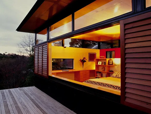 Exterior view of a compact modern timber-clad dwelling at dusk, photographed from a broad timber deck. Large floor-to-ceiling glazing reveals a warmly lit bedroom and built-in cabinetry inside, while dark framing, timber soffits, horizontal cladding, and operable louvred panels define the facade.