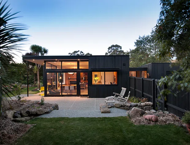 Exterior view of a compact, single-storey dark-clad home set within a landscaped garden. The building has a low-pitched or flat roof, extensive floor-to-ceiling glazing, timber-framed doors and windows, and a covered outdoor terrace with seating. Gravel paving, rock-edged garden beds, mature planting, and a dark privacy fence surround the house. Warm interior lighting creates an inviting evening atmosphere.