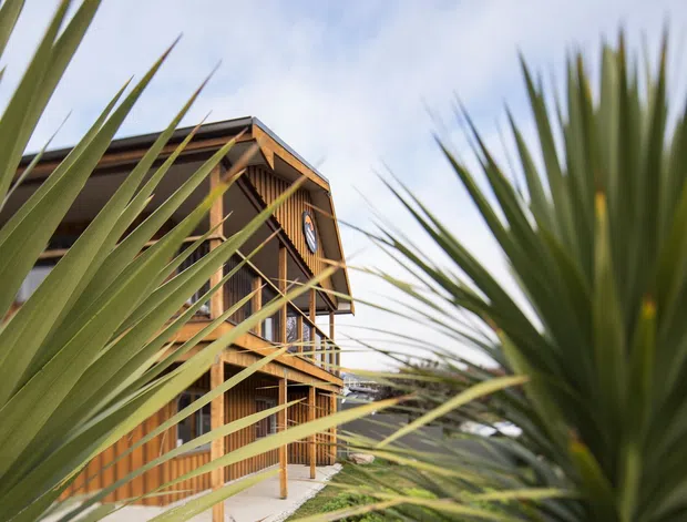 Exterior view of a multi-storey timber-clad building with prominent balconies, exposed structural posts and beams, and a pitched gable roof. Large spiky plants frame the foreground, with landscaped ground and a cloudy sky beyond.