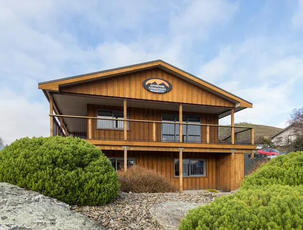 Exterior view of a two-storey timber-clad building with a prominent front-facing gable roof, deep eaves, and an upper-level balcony/verandah with simple timber posts and railings. The building sits among rocky landscaped terrain with low shrubs; a sign is mounted on the gable facade. A neighbouring house is partially visible in the background.