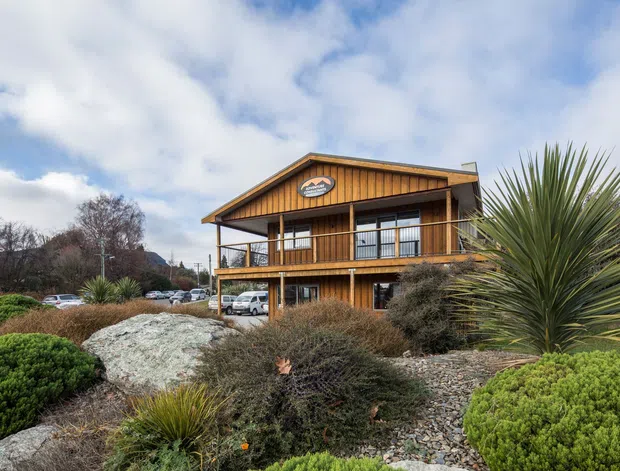 Exterior view of a two-storey timber-clad building with a prominent pitched gable roof, wraparound upper-level balcony, dark-framed glazing, and ground-level openings. The building is surrounded by extensive landscaped planting, gravel, rocks, and sculptural yucca-like vegetation. The setting appears semi-rural or suburban, with parked vehicles and mature trees in the background.