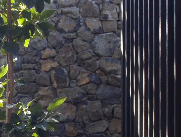 A close-up exterior architectural detail showing a dark vertical metal slatted screen beside a rough, irregular natural stone wall, framed by lush leafy planting. Strong sunlight creates alternating highlights and shadows across the screen and foliage.
