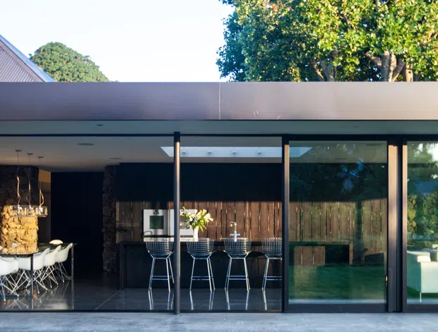 Contemporary single-storey house exterior with a long glazed façade beneath a broad flat roof. Through the floor-to-ceiling glazing, an open-plan kitchen and dining area with a central island, pendant lights, dining table and chairs is visible, along with a living room at the right. Dark-framed glazing, timber screening and stone-textured interior surfaces create a strong indoor-outdoor connection.