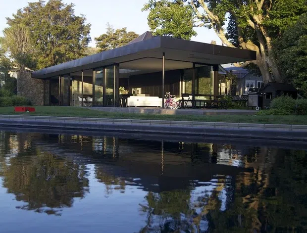 Exterior view of a low-slung modern pavilion beside a reflective swimming pool. The building has a broad, thin flat roof with deep overhangs, extensive floor-to-ceiling glazing, dark slender structural posts and frames, and an open-plan indoor-outdoor relationship. Mature trees surround the property, with a small portion of a neighbouring or secondary structure visible behind it.