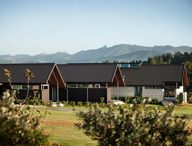 A low-rise contemporary rural building viewed from across landscaped grounds, featuring several connected gabled volumes with dark metal pitched roofs, dark cladding, timber-toned accents, and extensive horizontal glazing. Mountains and wooded hills form the background.
