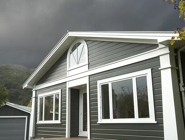 Exterior view of a compact single-storey gable-roof house with dark grey horizontal weatherboard cladding, crisp white architectural trims, white-framed windows, and a distinctive arched upper window. A detached garage is partly visible at the side, with a wooded hillside in the background.
