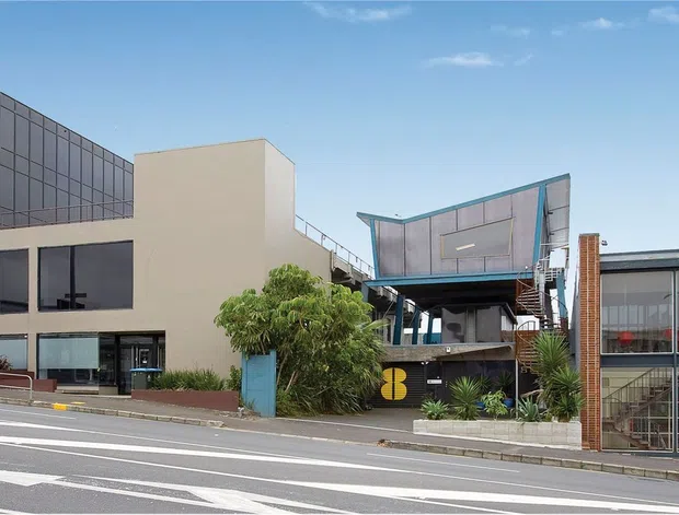 Street-level exterior view of a contemporary multi-storey commercial building complex with glazed office façades, beige rendered walls, a dark framed glass corner volume, and a smaller commercial frontage. The image shows a broad paved road in the foreground and urban streetscape context.