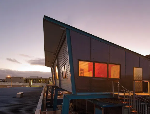 Exterior dusk view of a contemporary, angular multi-level building with a sharply sloping roofline, dark metal cladding, horizontal weatherboard-like siding, warm-lit windows, and an elevated deck or balcony with metal railings. The urban rooftop setting and dramatic evening lighting emphasize the building’s sculptural form.