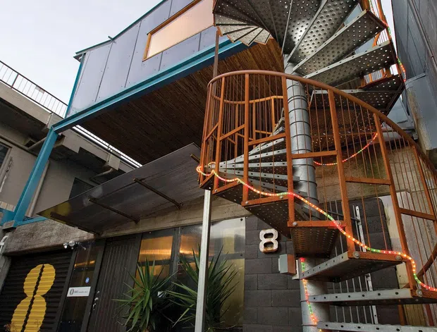 Exterior view of a contemporary multi-level commercial or mixed-use building, dominated by a sculptural metal spiral staircase with perforated steel treads and curved orange metal railings. The facade combines dark painted masonry, metal cladding, glazing, exposed structural elements and timber-like soffit lining. Potted plants and illuminated string lights sit near the entrance, which is marked with the number 8.