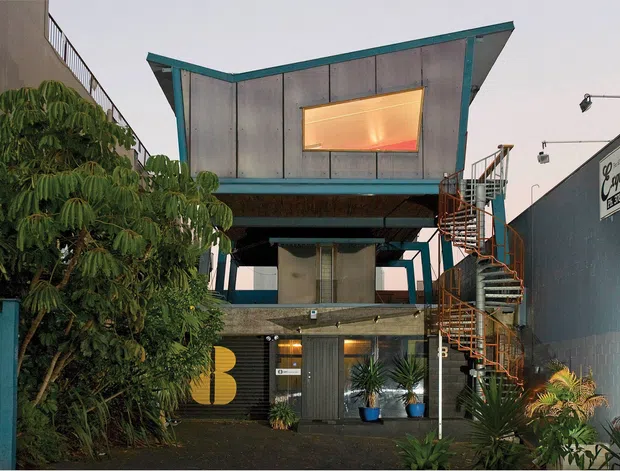 Exterior view of a compact multi-level commercial or mixed-use building with a distinctive cantilevered upper volume, corrugated metal cladding, teal-painted structural edges, an exposed spiral stair, and a glazed ground-floor frontage. Dense planting lines the narrow urban forecourt, and adjacent signage suggests a commercial setting.