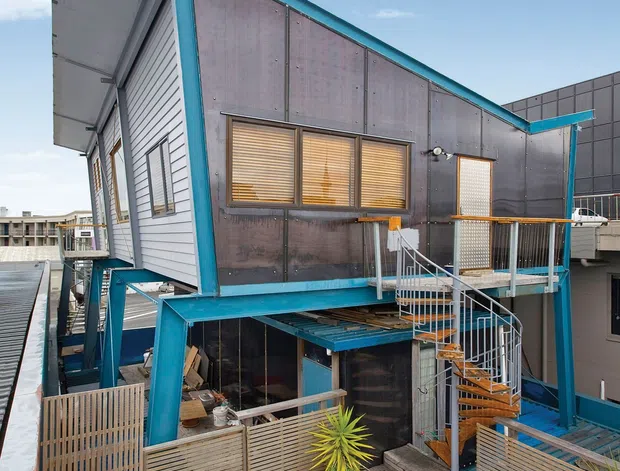 Exterior view of a distinctive multi-level urban building with a steeply sloping roofline, dark translucent-looking wall panels, pale horizontal cladding, and vivid turquoise structural framing and trim. The building is accessed by an external metal spiral staircase and surrounded by timber decking, slatted privacy fencing, and raised outdoor platforms.