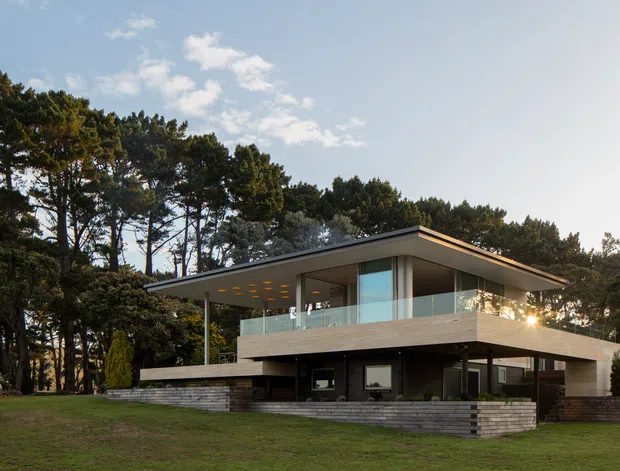 Exterior view of a contemporary two-storey flat-roof home set within a wooded landscape. The building features broad cantilevered roof planes, extensive floor-to-ceiling glazing, a glass-balustraded upper terrace, dark structural elements, and layered horizontal stone or concrete retaining walls.