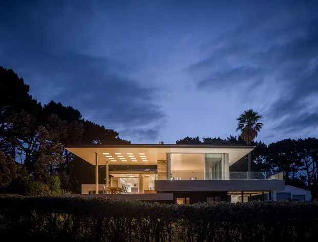 Twilight exterior photograph of a contemporary two-storey flat-roof house set within a landscaped, tree-lined site. The home has extensive glazing, warm illuminated interiors, projecting cantilevered volumes, a large sheltered outdoor terrace, and a dark-toned upper facade or balcony edge. A palm and mature trees frame the residence.