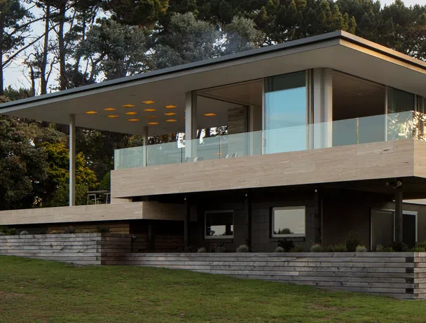 A contemporary two-storey flat-roof house set among mature trees, viewed from the front across a lawn. The building has a projecting upper level with broad glazing, a glass-balustraded terrace, a deep cantilevered roof, pale horizontal cladding, dark lower-level walls, and layered horizontal stone retaining walls.