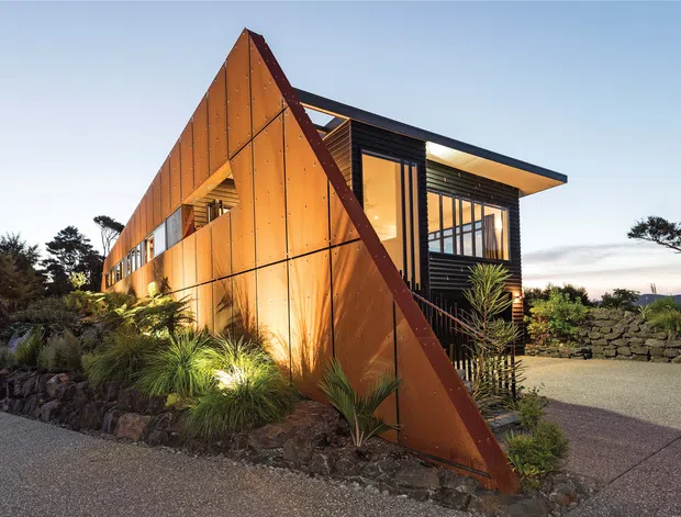 Exterior architectural photograph of a distinctive modern angular residence at dusk. The building features a sharply sloping triangular rust-coloured metal-clad wall, dark structural framing, extensive glazing, and a projecting flat roof. Native-style landscaped planting, stone retaining walls, and a paved driveway surround the house.