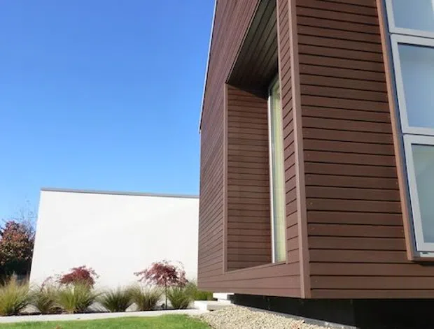 Exterior view of a contemporary single-storey building corner with dark horizontal timber cladding, deep recessed window openings, and large aluminium-framed glazing. The building sits above a dark base beside a gravel strip and neatly landscaped lawn under a clear blue sky.